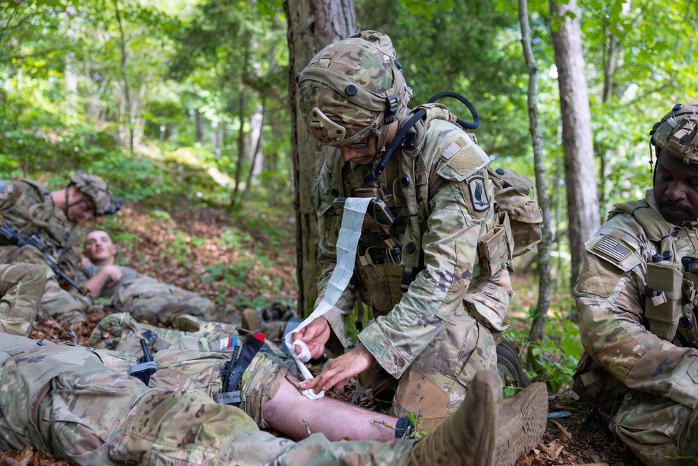 A military medical training exercise is taking place in a wooded outdoor environment. A soldier in full combat gear is applying bandaging to the leg of a casualty lying on the ground, while other soldiers are positioned nearby. The scene depicts tactical combat casualty care (TCCC) procedures being practiced or executed.