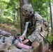 A military medical training exercise is taking place in a wooded outdoor environment. A soldier in full combat gear is applying bandaging to the leg of a casualty lying on the ground, while other soldiers are positioned nearby. The scene depicts tactical combat casualty care (TCCC) procedures being practiced or executed.