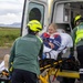 Two paramedics wearing green uniforms and yellow helmets labeled 'Sjúkrabíll Ambulance' are loading a patient on a stretcher into the back of an ambulance. The patient, a blonde woman, appears to have visible injuries including blood on her face and arm. The scene takes place outdoors in what appears to be an Icelandic landscape based on the Icelandic text on the uniforms.