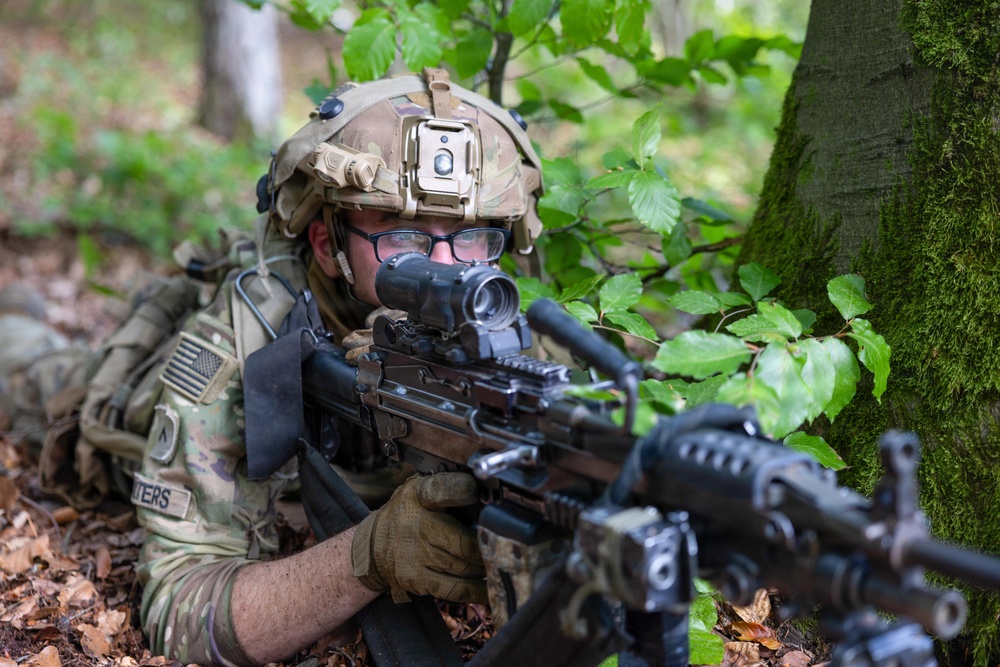 A U.S. Army soldier in full combat gear is positioned prone in a wooded area, aiming a rifle with an optical scope. The soldier is wearing MultiCam camouflage, a tactical helmet with a mounted device, and ballistic eyewear. The setting appears to be a training exercise or field operation in a deciduous forest environment.