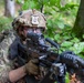 A U.S. Army soldier in full combat gear is positioned prone in a wooded area, aiming a rifle with an optical scope. The soldier is wearing MultiCam camouflage, a tactical helmet with a mounted device, and ballistic eyewear. The setting appears to be a training exercise or field operation in a deciduous forest environment.