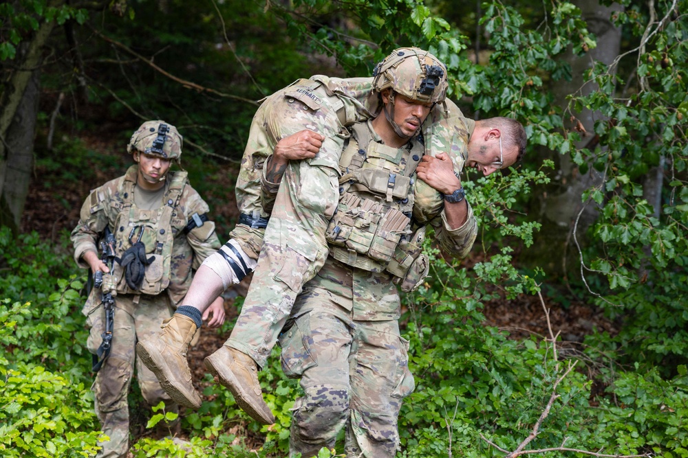 Military personnel in camouflage uniforms are conducting a casualty evacuation drill in a wooded outdoor environment. A soldier wearing a tactical helmet and plate carrier is performing a fireman's carry on another soldier who has simulated bandaging on their leg. A third soldier follows behind while carrying a rifle.