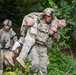 Military personnel in camouflage uniforms are conducting a casualty evacuation drill in a wooded outdoor environment. A soldier wearing a tactical helmet and plate carrier is performing a fireman's carry on another soldier who has simulated bandaging on their leg. A third soldier follows behind while carrying a rifle.