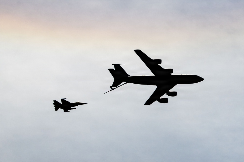 The image shows two military aircraft in silhouette against a pale, overcast sky at what appears to be dusk or dawn. A large tanker aircraft is extending a refueling boom toward a smaller fighter jet, suggesting an aerial refueling operation in progress. Both aircraft are rendered as dark silhouettes, providing no color or markings detail.