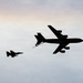 The image shows two military aircraft in silhouette against a pale, overcast sky at what appears to be dusk or dawn. A large tanker aircraft is extending a refueling boom toward a smaller fighter jet, suggesting an aerial refueling operation in progress. Both aircraft are rendered as dark silhouettes, providing no color or markings detail.