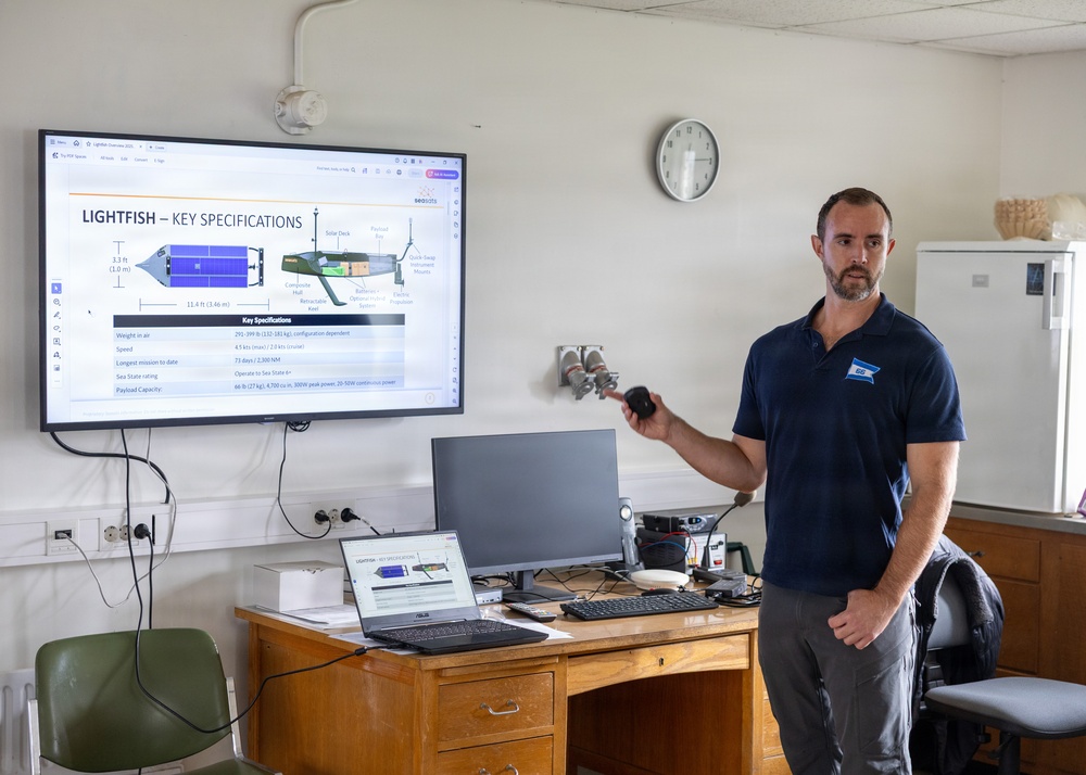 A man in a navy polo shirt is presenting a technical briefing about a maritime autonomous vehicle called 'Lightfish' in what appears to be a classroom or office setting. He is standing next to a large wall-mounted display showing the 'Lightfish – Key Specifications' slide, while holding a presentation remote. A desk in front of him holds a laptop, monitor, keyboard, and various equipment.