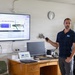 A man in a navy polo shirt is presenting a technical briefing about a maritime autonomous vehicle called 'Lightfish' in what appears to be a classroom or office setting. He is standing next to a large wall-mounted display showing the 'Lightfish – Key Specifications' slide, while holding a presentation remote. A desk in front of him holds a laptop, monitor, keyboard, and various equipment.