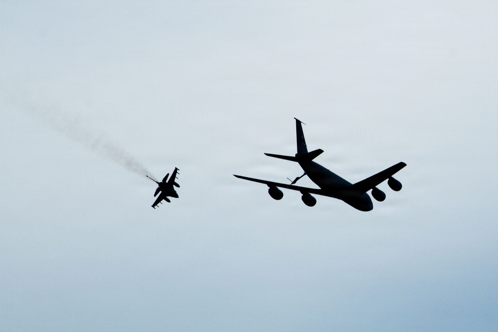 Two military aircraft are captured in silhouette against an overcast sky. A large multi-engine tanker or transport aircraft flies in close proximity to a smaller single-engine fighter jet. The fighter jet appears to be trailing a visible smoke or exhaust plume.