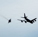 Two military aircraft are captured in silhouette against an overcast sky. A large multi-engine tanker or transport aircraft flies in close proximity to a smaller single-engine fighter jet. The fighter jet appears to be trailing a visible smoke or exhaust plume.