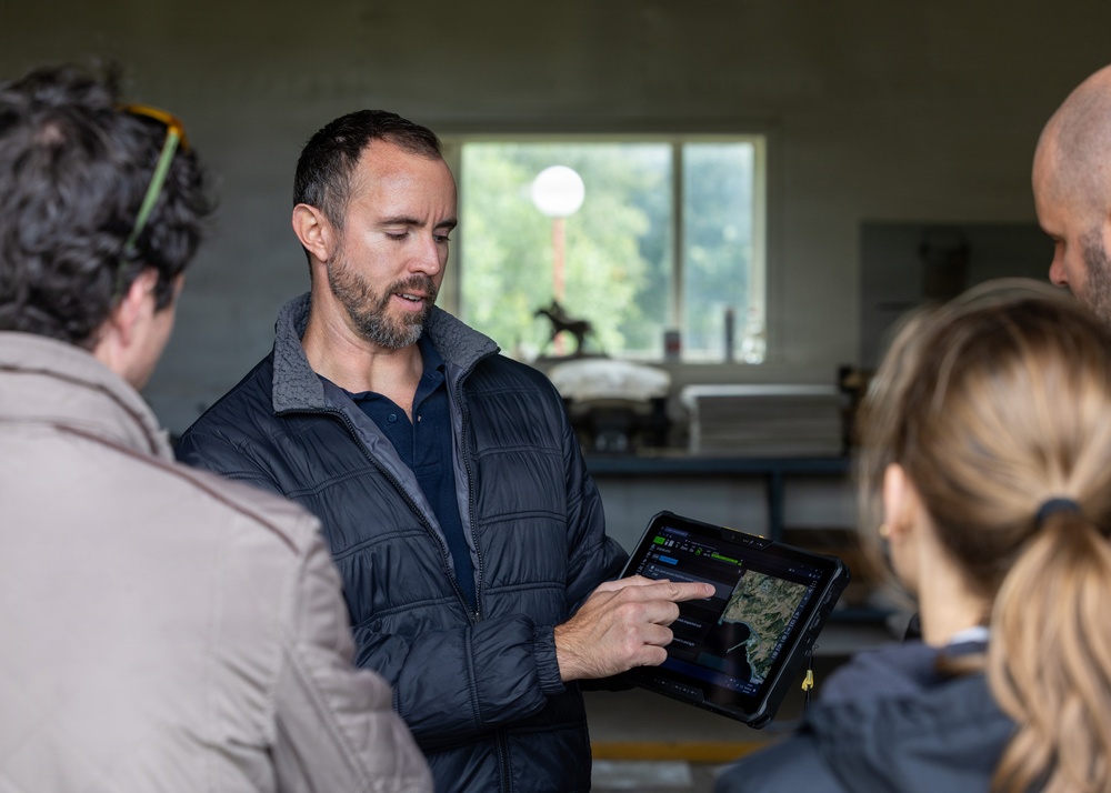 A man wearing a dark quilted vest is presenting information on a rugged tablet device to a small group of people in an indoor setting. The tablet appears to display a satellite or aerial map with a dark-themed interface. The setting appears to be a casual workspace or outbuilding with a window visible in the background.