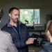 A man wearing a dark quilted vest is presenting information on a rugged tablet device to a small group of people in an indoor setting. The tablet appears to display a satellite or aerial map with a dark-themed interface. The setting appears to be a casual workspace or outbuilding with a window visible in the background.