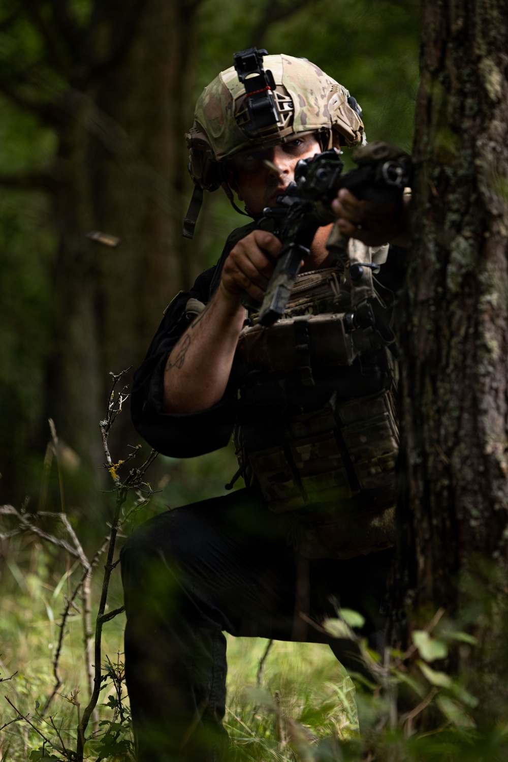 A soldier in full tactical gear is positioned behind a tree in a forested environment, aiming a rifle with an optic sight. The individual is wearing a multicam helmet with a night vision mount, plate carrier vest, and dark clothing. A spent brass casing appears to be ejecting from the weapon, suggesting active firing.