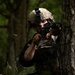 A soldier in full tactical gear is positioned behind a tree in a forested environment, aiming a rifle with an optic sight. The individual is wearing a multicam helmet with a night vision mount, plate carrier vest, and dark clothing. A spent brass casing appears to be ejecting from the weapon, suggesting active firing.