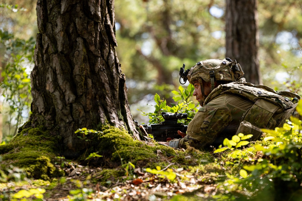 A soldier in full combat gear is lying prone on a moss-covered forest floor, taking up a defensive or observation position beside a large tree trunk. The soldier is equipped with a tactical helmet, body armor, backpack, and is aiming or holding a weapon with an optical sight. The image appears to depict a military field exercise or training scenario in a deciduous/mixed woodland environment.