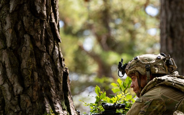A soldier in full combat gear is lying prone on a moss-covered forest floor, taking up a defensive or observation position beside a large tree trunk. The soldier is equipped with a tactical helmet, body armor, backpack, and is aiming or holding a weapon with an optical sight. The image appears to depict a military field exercise or training scenario in a deciduous/mixed woodland environment.