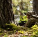 A soldier in full combat gear is lying prone on a moss-covered forest floor, taking up a defensive or observation position beside a large tree trunk. The soldier is equipped with a tactical helmet, body armor, backpack, and is aiming or holding a weapon with an optical sight. The image appears to depict a military field exercise or training scenario in a deciduous/mixed woodland environment.