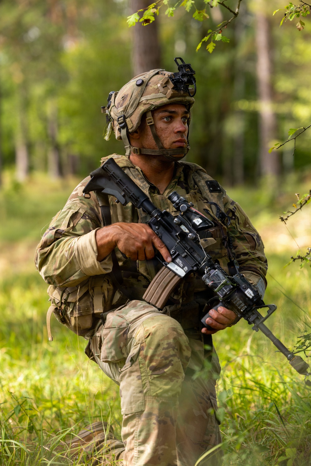 A soldier in full combat gear is positioned in a wooded, grassy area while holding an assault rifle at the ready. The soldier is wearing multicam camouflage uniform, a tactical helmet with night vision mount, and a full kit including body armor and rucksack. The image appears to be taken during a field training exercise or operational patrol.