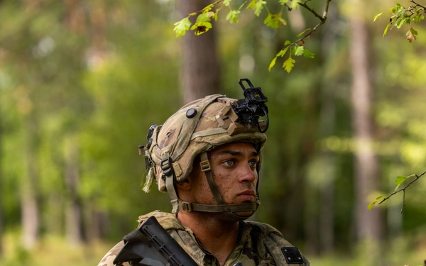 A soldier in full combat gear is positioned in a wooded, grassy area while holding an assault rifle at the ready. The soldier is wearing multicam camouflage uniform, a tactical helmet with night vision mount, and a full kit including body armor and rucksack. The image appears to be taken during a field training exercise or operational patrol.