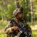 A soldier in full combat gear is positioned in a wooded, grassy area while holding an assault rifle at the ready. The soldier is wearing multicam camouflage uniform, a tactical helmet with night vision mount, and a full kit including body armor and rucksack. The image appears to be taken during a field training exercise or operational patrol.