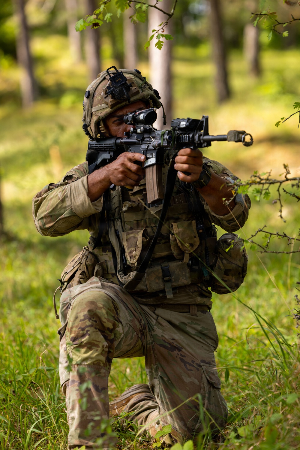 A soldier in full combat gear is kneeling in a wooded, grassy area while aiming a rifle with an optical scope. The soldier is wearing MultiCam/OCP camouflage uniform, a tactical helmet with night vision mount, and a plate carrier vest with magazine pouches. The image appears to depict a military training exercise or field operation.