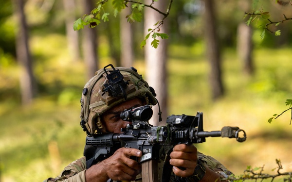 A soldier in full combat gear is kneeling in a wooded, grassy area while aiming a rifle with an optical scope. The soldier is wearing MultiCam/OCP camouflage uniform, a tactical helmet with night vision mount, and a plate carrier vest with magazine pouches. The image appears to depict a military training exercise or field operation.