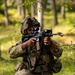 A soldier in full combat gear is kneeling in a wooded, grassy area while aiming a rifle with an optical scope. The soldier is wearing MultiCam/OCP camouflage uniform, a tactical helmet with night vision mount, and a plate carrier vest with magazine pouches. The image appears to depict a military training exercise or field operation.