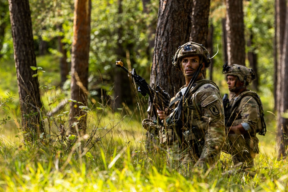 Two soldiers in full combat gear are positioned in a forested area with tall grass and pine trees. The foreground soldier is holding a rifle with a training blank-firing adapter attached to the muzzle and is looking directly at the camera. The scene appears to be a military field training exercise based on the equipment and environment.