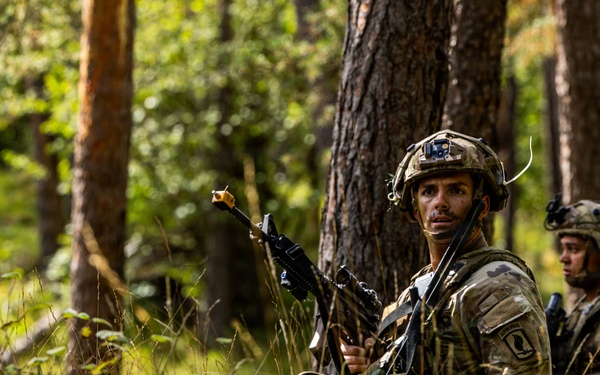 Two soldiers in full combat gear are positioned in a forested area with tall grass and pine trees. The foreground soldier is holding a rifle with a training blank-firing adapter attached to the muzzle and is looking directly at the camera. The scene appears to be a military field training exercise based on the equipment and environment.