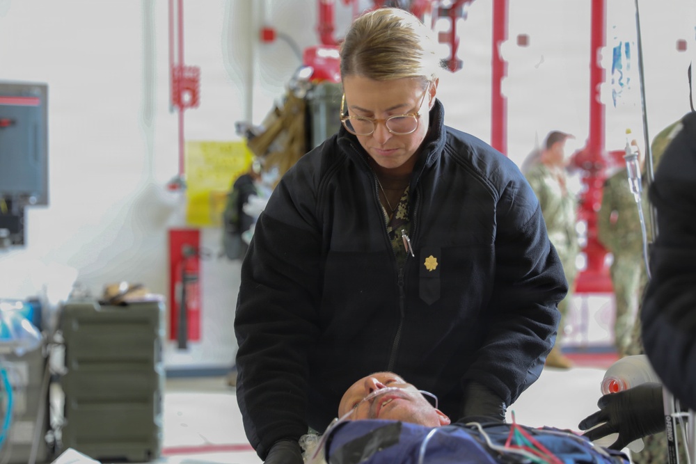 A medical professional wearing a black jacket with a gold oak leaf insignia is attending to a patient lying on a stretcher or gurney in what appears to be a military medical facility or hangar. The provider is leaning over the patient, who has medical leads or tubes attached, suggesting active patient assessment or treatment. Military personnel in camouflage uniforms are visible in the background.