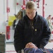 A medical professional wearing a black jacket with a gold oak leaf insignia is attending to a patient lying on a stretcher or gurney in what appears to be a military medical facility or hangar. The provider is leaning over the patient, who has medical leads or tubes attached, suggesting active patient assessment or treatment. Military personnel in camouflage uniforms are visible in the background.