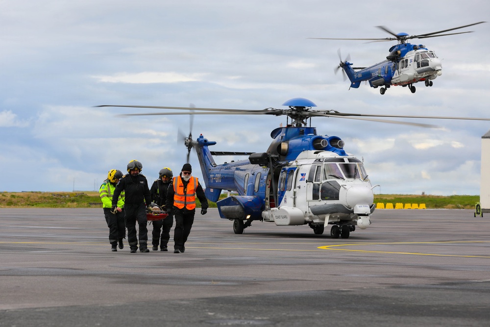 A search and rescue operation is underway on a tarmac apron, with personnel carrying a stretcher with a patient away from a grounded blue-and-white Coast Guard helicopter. A second identical helicopter is airborne in the background. The scene appears to be at a coastal airfield, likely in Iceland based on the visible registration markings 'TF-GRO' on the grounded aircraft.