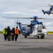 A search and rescue operation is underway on a tarmac apron, with personnel carrying a stretcher with a patient away from a grounded blue-and-white Coast Guard helicopter. A second identical helicopter is airborne in the background. The scene appears to be at a coastal airfield, likely in Iceland based on the visible registration markings 'TF-GRO' on the grounded aircraft.