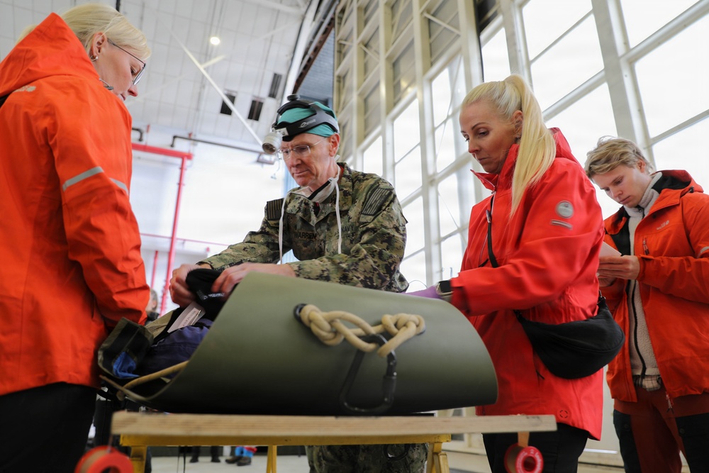 A military personnel member in U.S. Navy camouflage uniform, wearing goggles on his head, demonstrates or examines equipment on a table alongside several civilians wearing orange/red jackets. The setting appears to be a large hangar or industrial facility with large windows in the background. The group appears to be engaged in a training, demonstration, or briefing activity involving what appears to be a rolled military-grade mat or stretcher with rope.