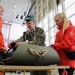 A military personnel member in U.S. Navy camouflage uniform, wearing goggles on his head, demonstrates or examines equipment on a table alongside several civilians wearing orange/red jackets. The setting appears to be a large hangar or industrial facility with large windows in the background. The group appears to be engaged in a training, demonstration, or briefing activity involving what appears to be a rolled military-grade mat or stretcher with rope.