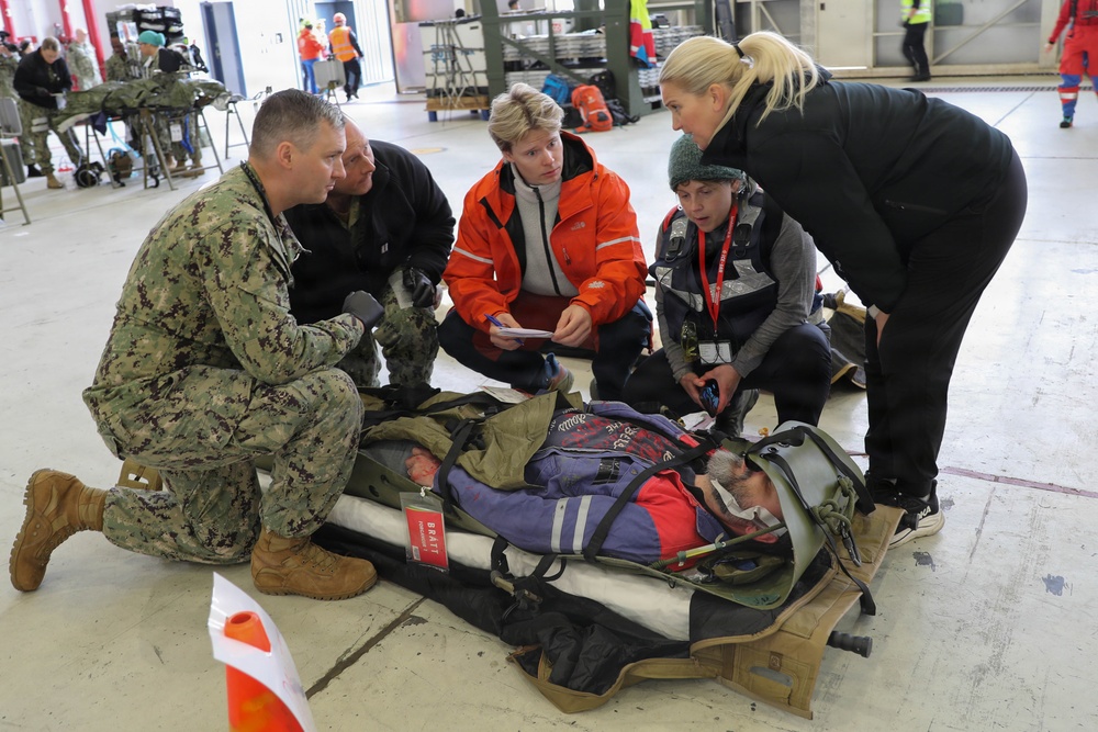 A multi-agency emergency response exercise or drill is taking place inside what appears to be a large hangar or warehouse facility. A person on a stretcher/litter is being assessed by a group including a US Navy sailor in digital camouflage uniform, civilian responders, and medical personnel. The scene suggests a joint military-civilian mass casualty training exercise.