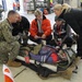 A multi-agency emergency response exercise or drill is taking place inside what appears to be a large hangar or warehouse facility. A person on a stretcher/litter is being assessed by a group including a US Navy sailor in digital camouflage uniform, civilian responders, and medical personnel. The scene suggests a joint military-civilian mass casualty training exercise.