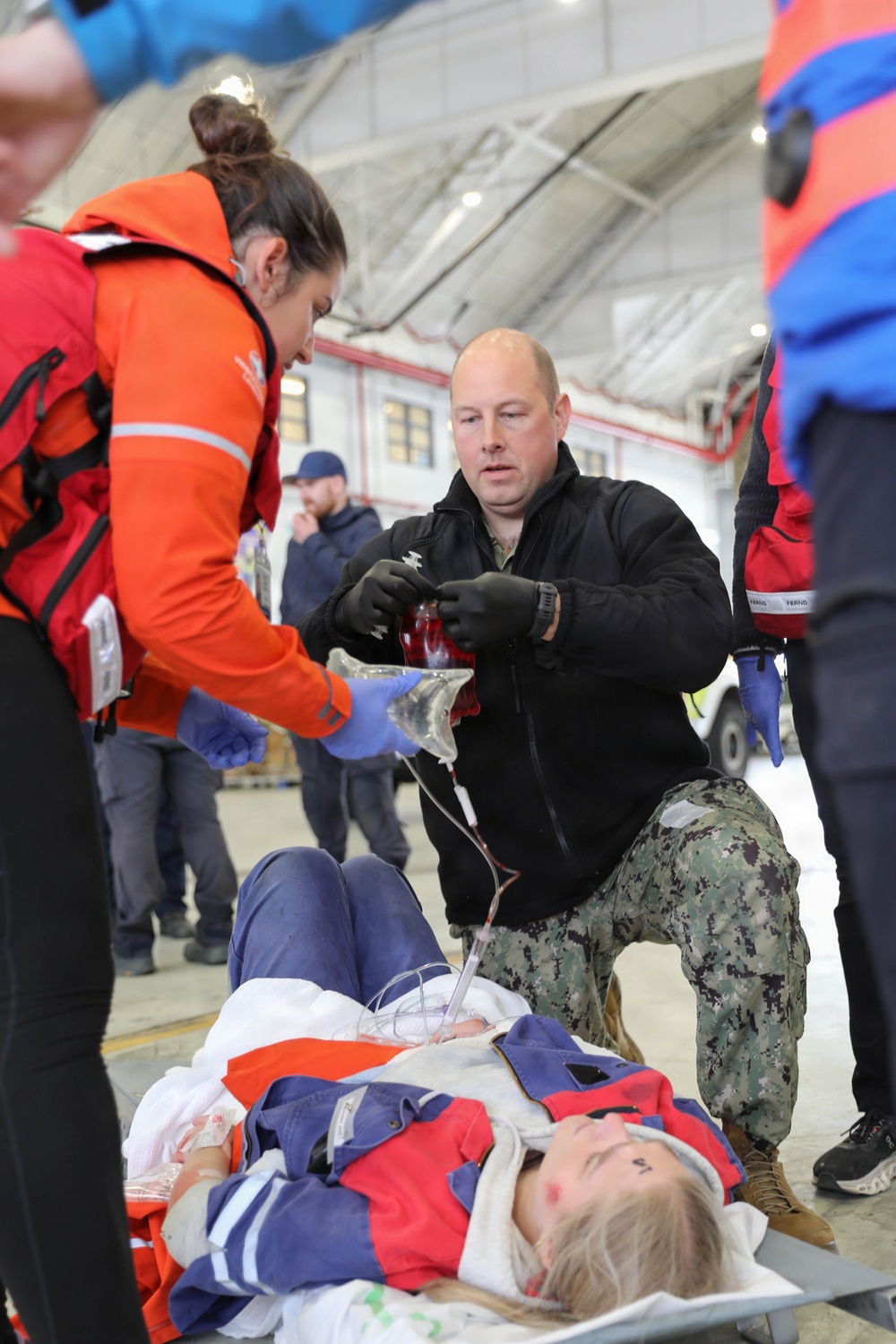 A medical response scene inside what appears to be a hangar or large indoor facility, where personnel are treating a patient lying on a stretcher. A person in US Navy camouflage pants and black fleece is administering IV fluids while a person in an orange rescue/survival suit assists. The scene appears to be either a real emergency response or a training exercise.