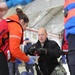 A medical response scene inside what appears to be a hangar or large indoor facility, where personnel are treating a patient lying on a stretcher. A person in US Navy camouflage pants and black fleece is administering IV fluids while a person in an orange rescue/survival suit assists. The scene appears to be either a real emergency response or a training exercise.