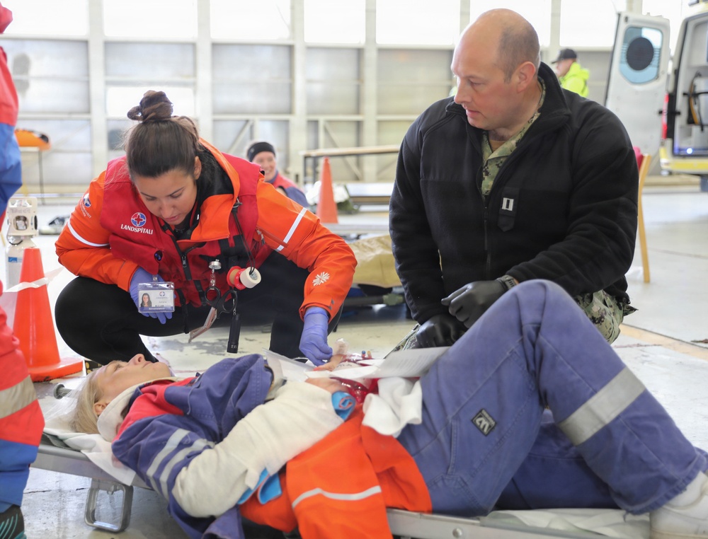 A medical training or emergency drill exercise is taking place inside what appears to be a hangar or large indoor facility. A female medical responder wearing an orange 'Landspitali' jacket and blue gloves is attending to a simulated casualty lying on a stretcher, while a military officer in a black fleece with rank insignia observes. The scene includes simulated injuries, medical equipment, and multiple personnel in the background.
