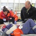 A medical training or emergency drill exercise is taking place inside what appears to be a hangar or large indoor facility. A female medical responder wearing an orange 'Landspitali' jacket and blue gloves is attending to a simulated casualty lying on a stretcher, while a military officer in a black fleece with rank insignia observes. The scene includes simulated injuries, medical equipment, and multiple personnel in the background.
