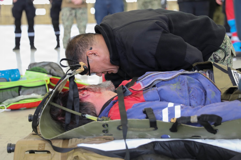 A responder in a black hoodie is leaning over a person lying on a military-style litter/stretcher, appearing to assess or perform airway/breathing checks. The person on the stretcher has visible simulated blood/wounds on their head and is wearing a red and blue jacket with reflective striping. This appears to be a medical training exercise or drill, with additional personnel and equipment visible in the background.
