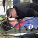 A responder in a black hoodie is leaning over a person lying on a military-style litter/stretcher, appearing to assess or perform airway/breathing checks. The person on the stretcher has visible simulated blood/wounds on their head and is wearing a red and blue jacket with reflective striping. This appears to be a medical training exercise or drill, with additional personnel and equipment visible in the background.