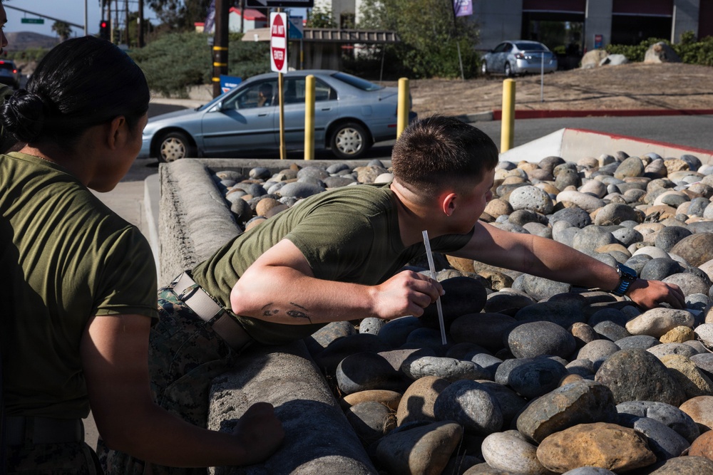 Two individuals wearing olive green military-style uniforms are near a decorative rock/boulder landscaping feature along a roadway. One person is leaning over the rock barrier holding what appears to be a straw or thin tube, while the other stands observing. The setting appears to be a commercial or base entrance area with vehicles, bollards, and signage visible in the background.