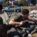 Two individuals wearing olive green military-style uniforms are near a decorative rock/boulder landscaping feature along a roadway. One person is leaning over the rock barrier holding what appears to be a straw or thin tube, while the other stands observing. The setting appears to be a commercial or base entrance area with vehicles, bollards, and signage visible in the background.