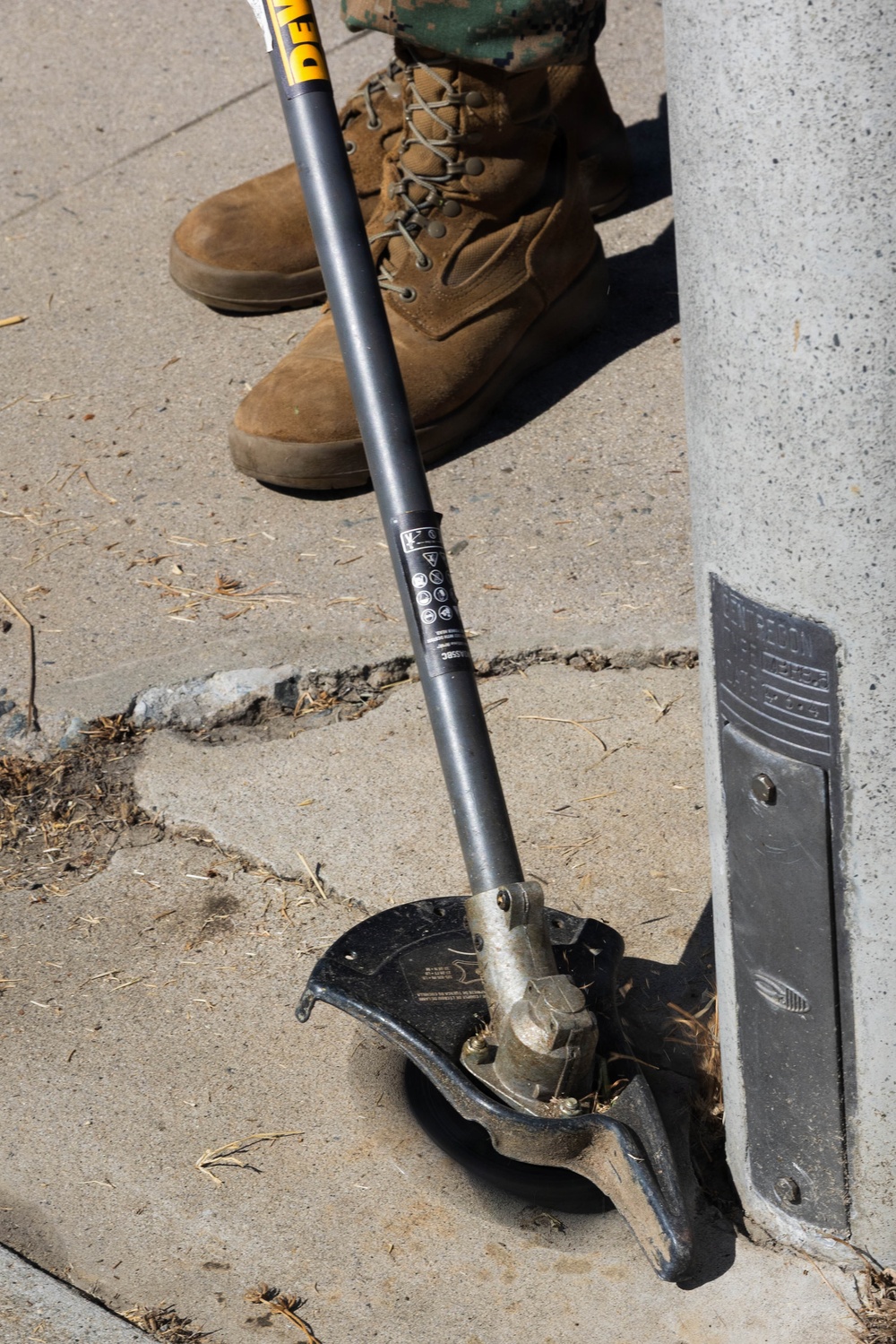 A DeWalt string trimmer/weed eater is resting against a concrete pillar or post on a sidewalk. The cutting head is visible at the bottom, resting on the concrete surface with debris around it. A person wearing military-style combat boots and camouflage pants stands nearby in the background.