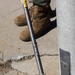 A DeWalt string trimmer/weed eater is resting against a concrete pillar or post on a sidewalk. The cutting head is visible at the bottom, resting on the concrete surface with debris around it. A person wearing military-style combat boots and camouflage pants stands nearby in the background.