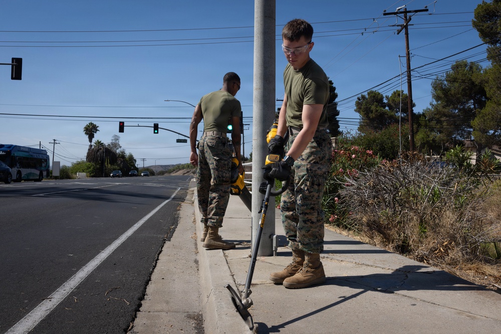 Two military personnel in camouflage uniforms and olive drab t-shirts are performing roadside maintenance work on a sidewalk along a suburban road. The foreground individual is using a DeWalt-branded power tool, likely a string trimmer or edger, near a utility pole while wearing safety glasses and gloves. The scene appears to be a community service or base beautification effort in a sunny, suburban environment.
