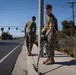 Two military personnel in camouflage uniforms and olive drab t-shirts are performing roadside maintenance work on a sidewalk along a suburban road. The foreground individual is using a DeWalt-branded power tool, likely a string trimmer or edger, near a utility pole while wearing safety glasses and gloves. The scene appears to be a community service or base beautification effort in a sunny, suburban environment.