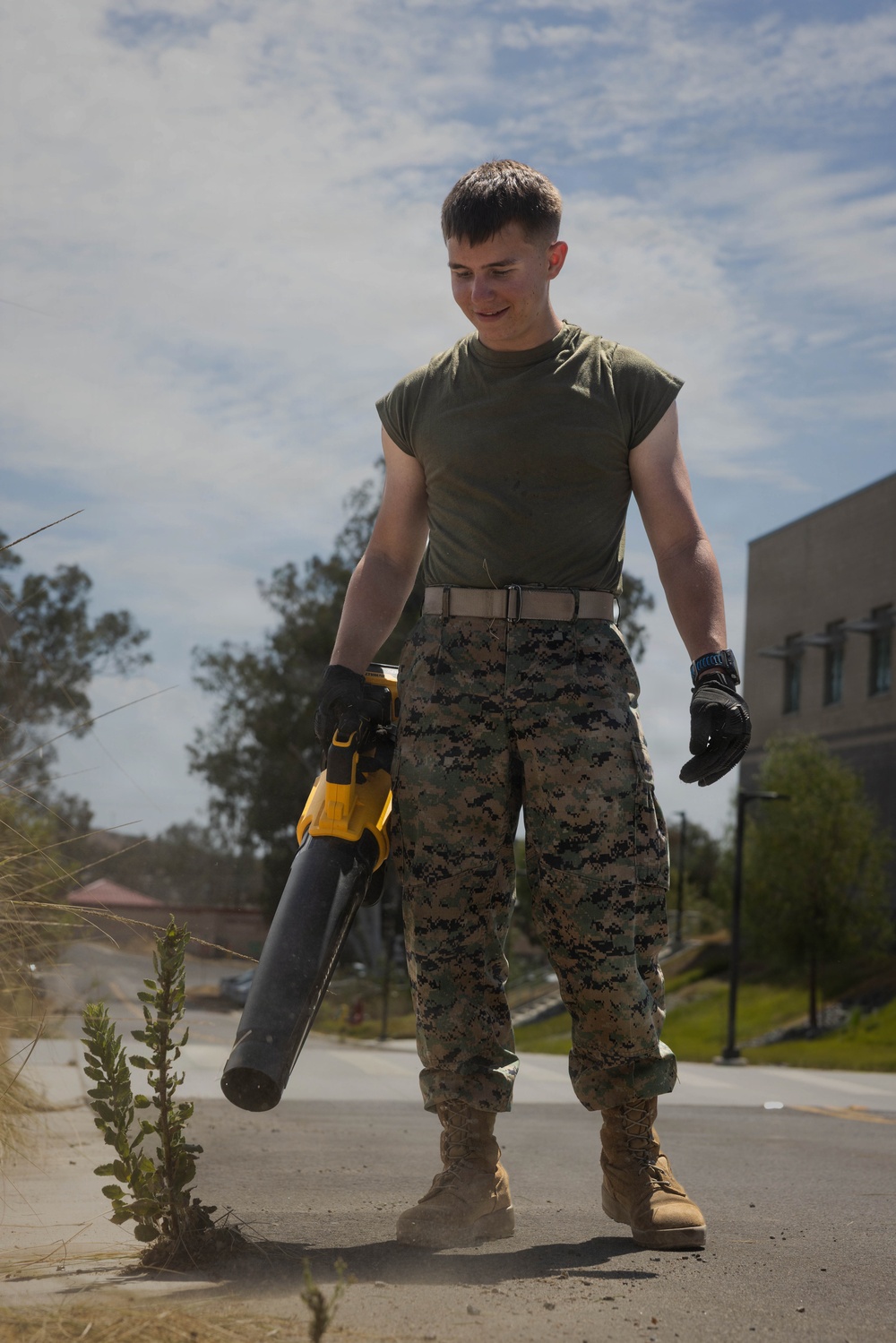 A young male service member wearing U.S. Marine Corps MARPAT digital camouflage trousers and an olive drab t-shirt is performing grounds maintenance on a paved surface. He is holding a yellow and black DeWalt cordless leaf blower and wearing tactical gloves and tan combat boots. The setting appears to be a military installation based on the uniform and background buildings.