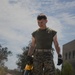 A young male service member wearing U.S. Marine Corps MARPAT digital camouflage trousers and an olive drab t-shirt is performing grounds maintenance on a paved surface. He is holding a yellow and black DeWalt cordless leaf blower and wearing tactical gloves and tan combat boots. The setting appears to be a military installation based on the uniform and background buildings.